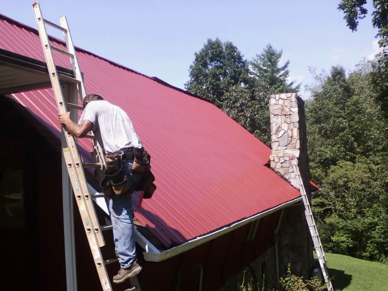 Roofer on a ladder beside a red metal roof and stone chimney — Soffit & Fascia Repair by Roofing Brothers in Pacific City